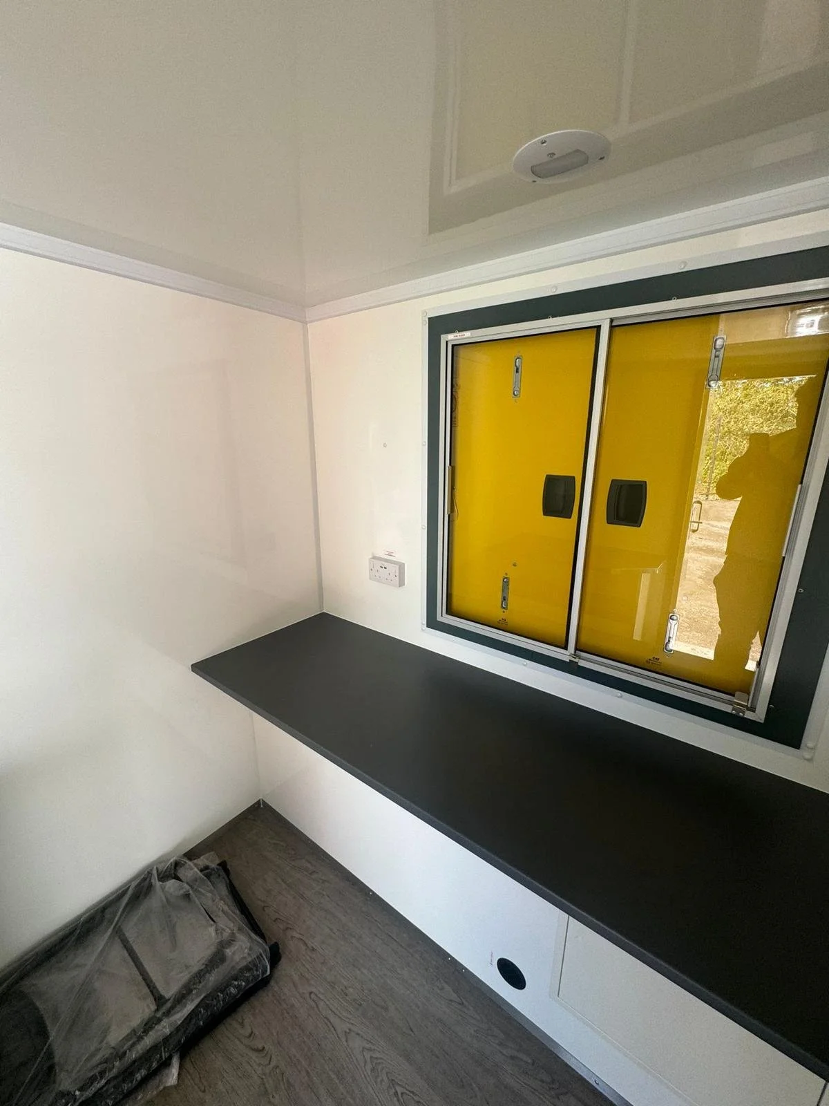 A small room with white walls, a dark countertop, and a yellow window with black sliding handles. There's a plastic-covered object on the floor in the corner and a ceiling-mounted smoke detector.