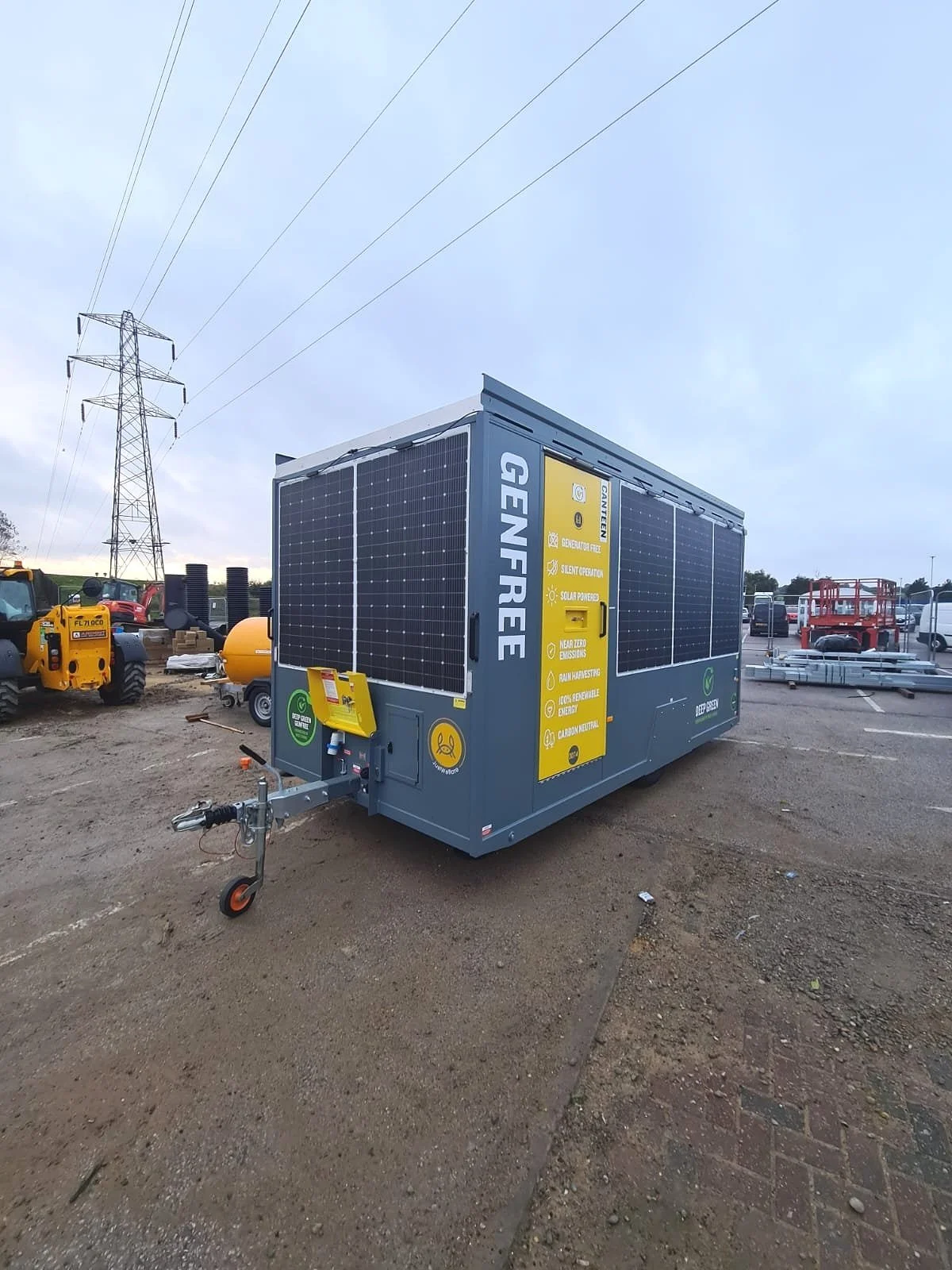 Portable generator with solar panels installed, labeled 'GENFREE,' situated on a dirt lot with construction equipment and power lines in the background.