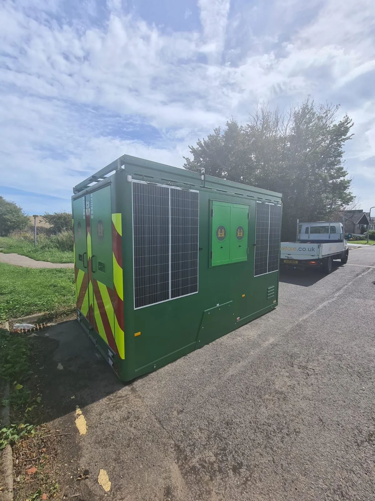Green electrical utility box with solar panels on its sides, located on the side of a paved road with a grassy area and trees in the background.
