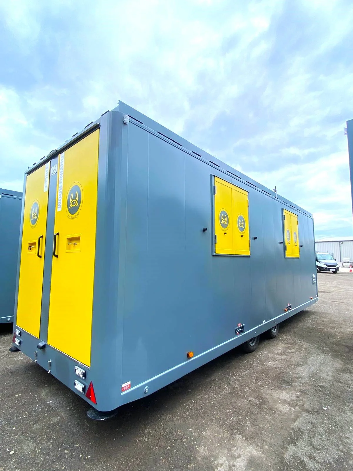 A portable restroom or toilet trailer with yellow doors and windows, mounted on wheels, outdoors on a dirt lot under a cloudy sky.