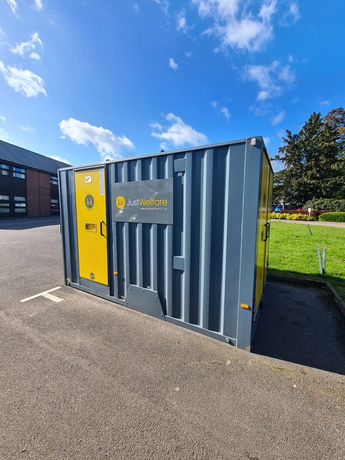 A grey and yellow mobile welfare unit with a logo and website for Just Welfare, parked in a parking lot with a building, trees, and a blue sky with clouds in the background.