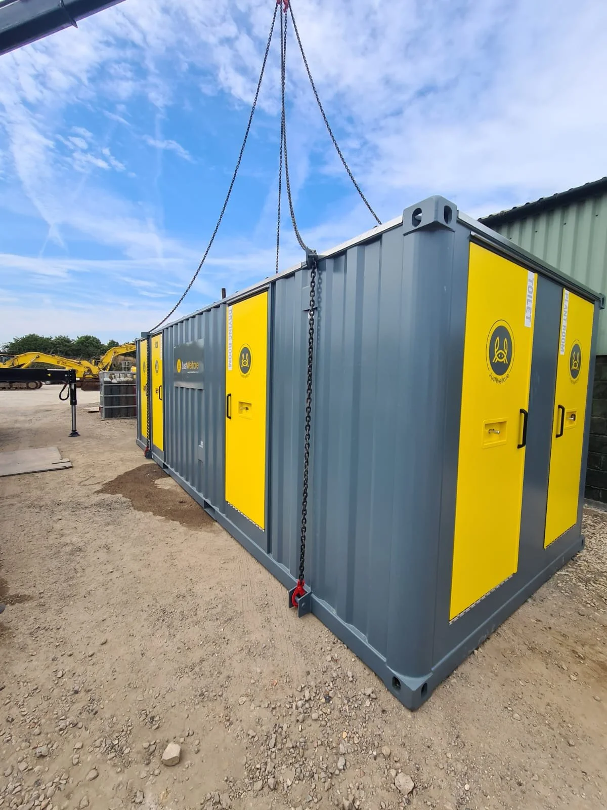 A gray and yellow portable generator container is being lifted with chains at an outdoor construction site.