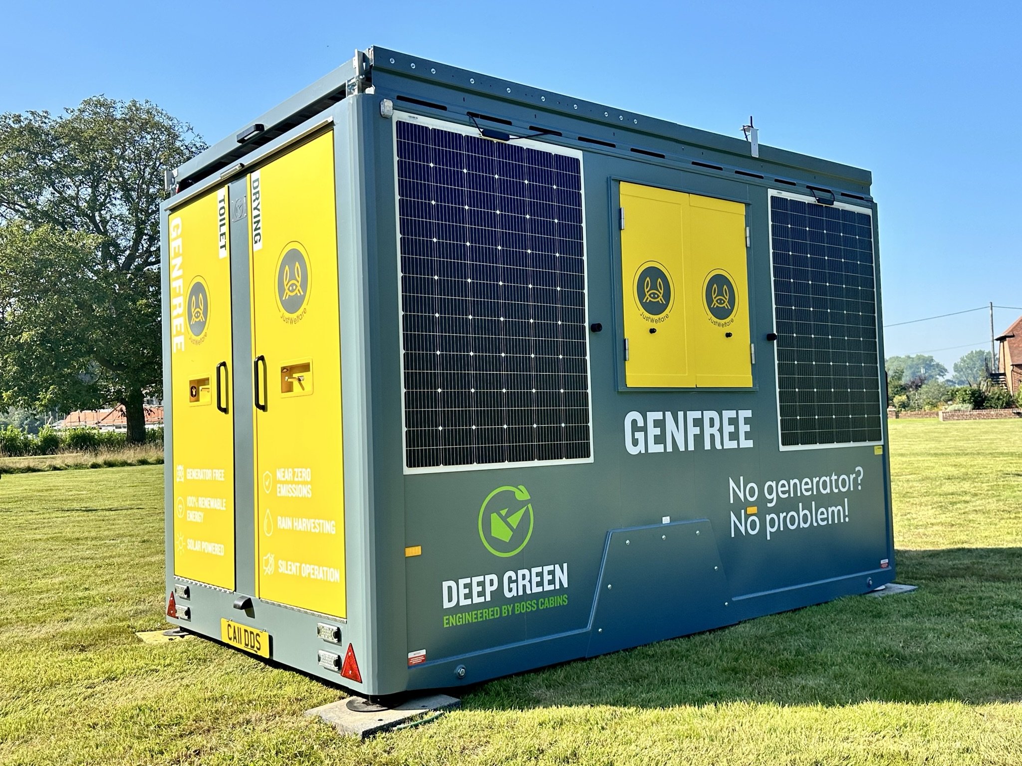 A portable generator with solar panels, labeled 'Genfree,' positioned on a grassy field against a backdrop of trees and a clear blue sky. The generator has yellow doors with icons, and text indicating it is environmentally friendly, renewable, and silent.