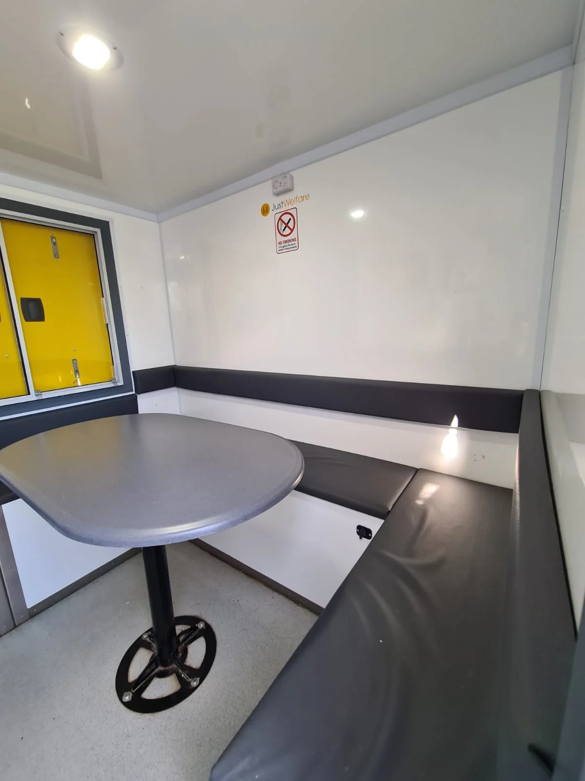 Small enclosed seating area with a gray table and black cushioned bench seating along the walls. There is a window with yellow shutters, a no smoking sign, and a whiteboard on the wall.