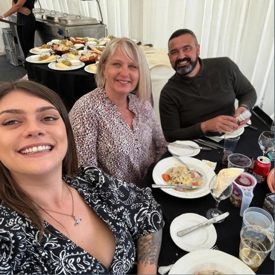 Three people sitting at a table enjoying a meal with plates of food, drinks, and condiments on the table. In the background, a buffet table with various dishes is visible.