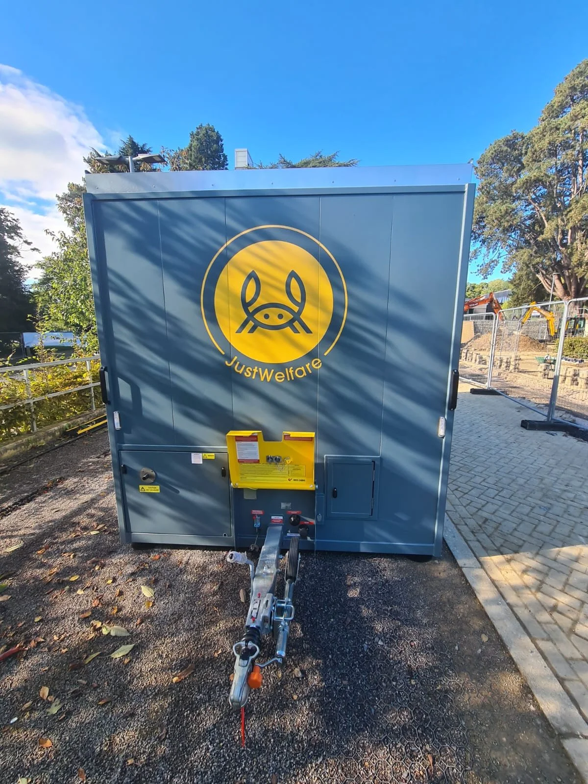 A gray trailer with a large yellow logo of a crab and the words 'Just Welfare' on it, parked outdoors with trees and construction equipment visible in the background.