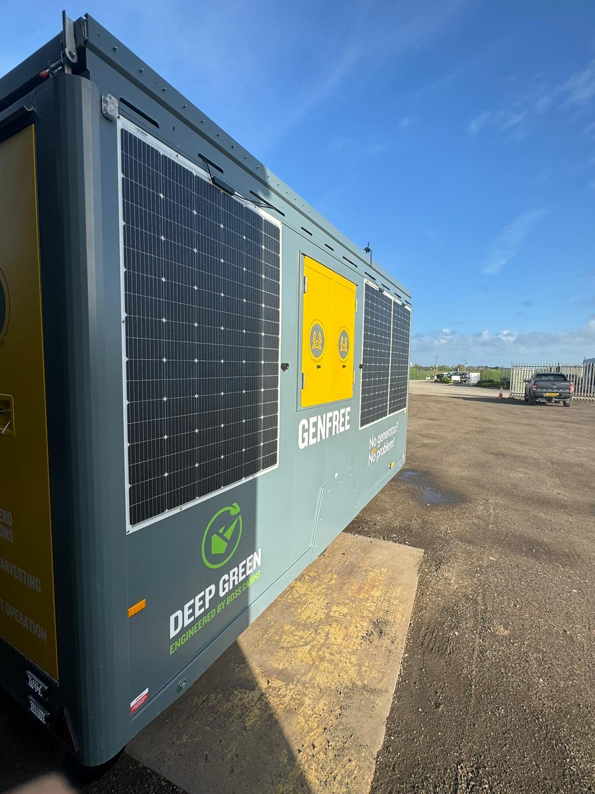A large gray generator with solar panels attached to its sides, labeled 'Deep Green' and 'GenFree,' located in an outdoor parking lot under a blue sky with some clouds.