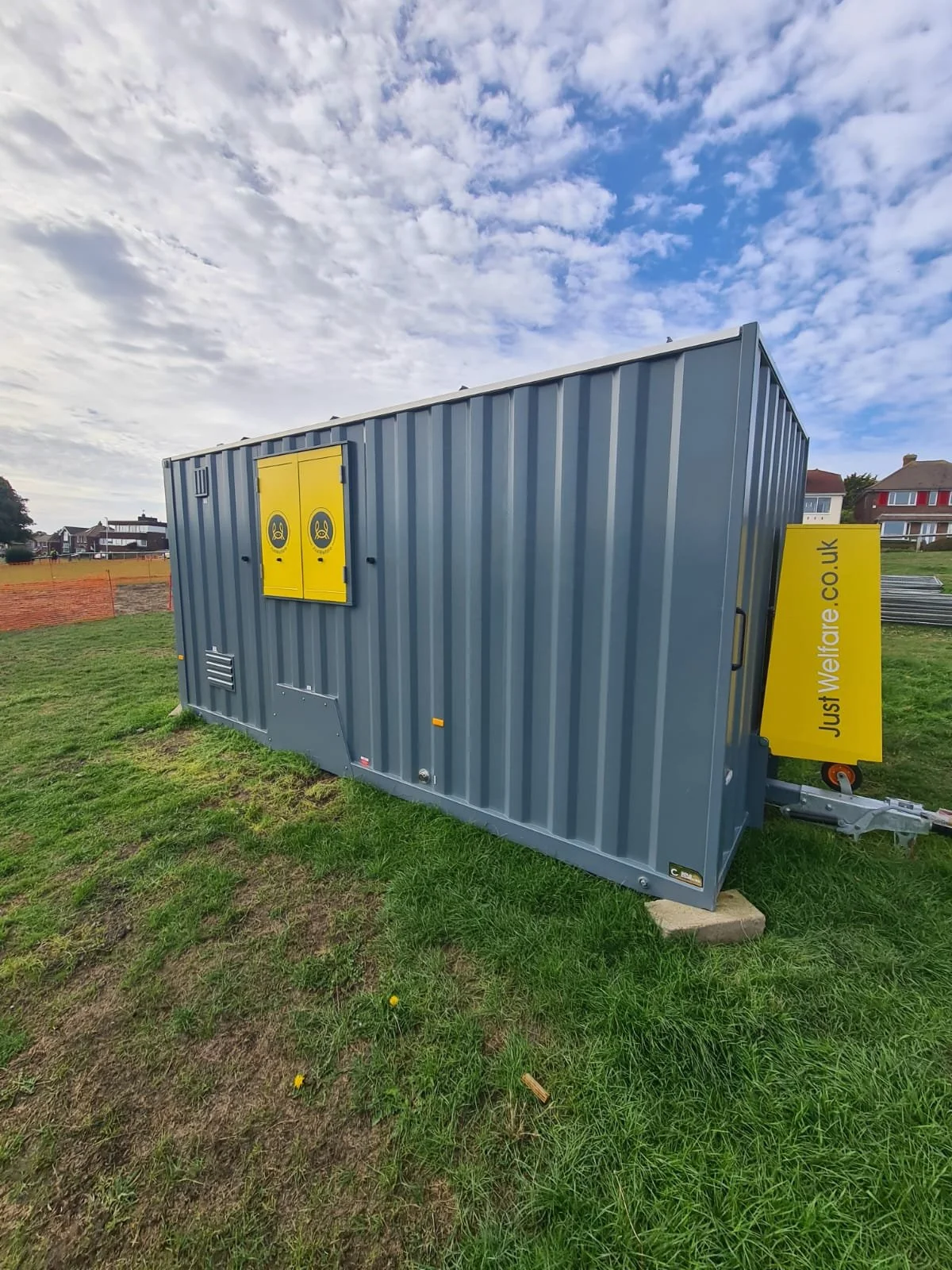 Gray mobile restroom trailer with yellow accents and biohazard symbols, situated on green grass under a partly cloudy sky.