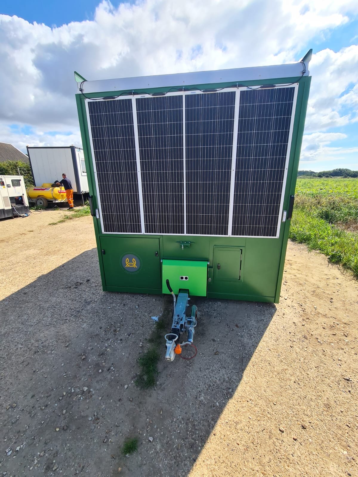 Green mobile generator or monitoring unit with solar panels on top, situated outdoors on dirt ground, with a shadow cast to the right and cloudy blue sky above.