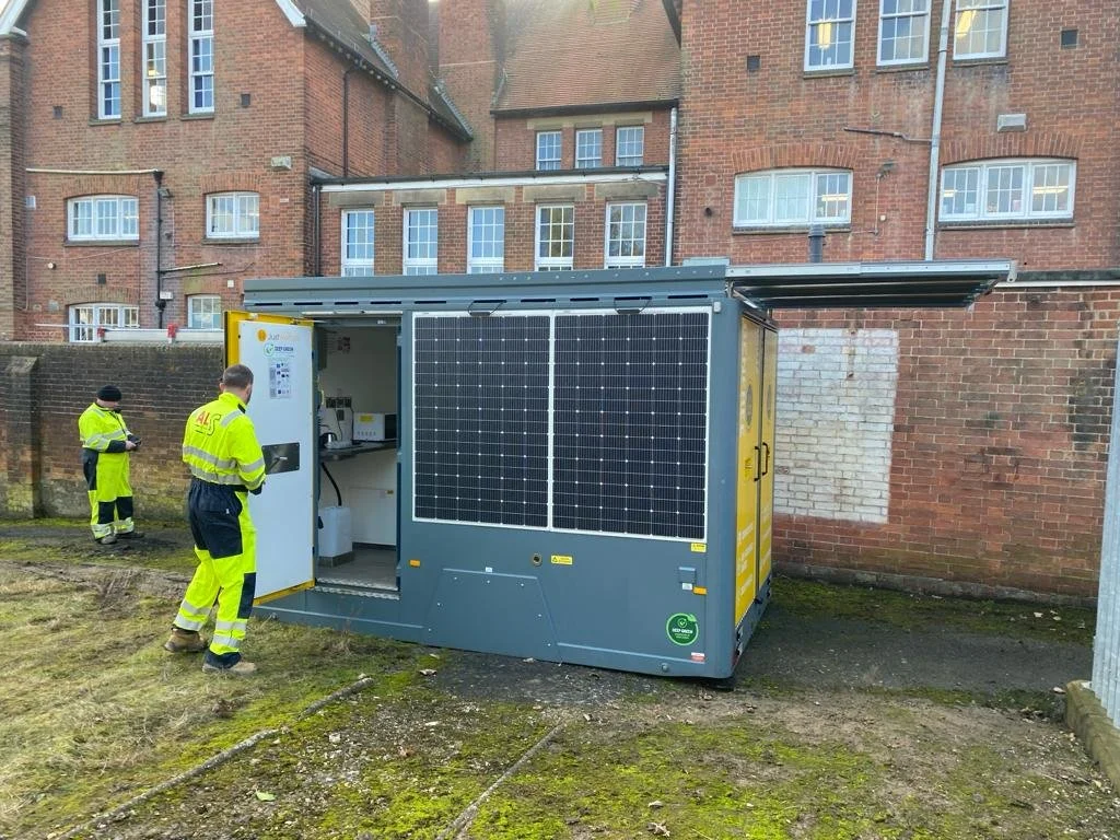 Two workers in yellow safety vests and black pants working near a portable generator with solar panels, beside a brick building.