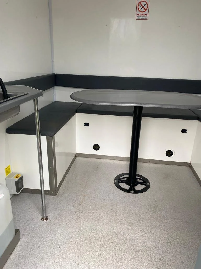 Empty corner of a room with a small round table, corner seating with black cushions, and a sink with a countertop on the left. There is a 'No Smoking' sign on the wall and ventilation holes on the lower part of the wall.