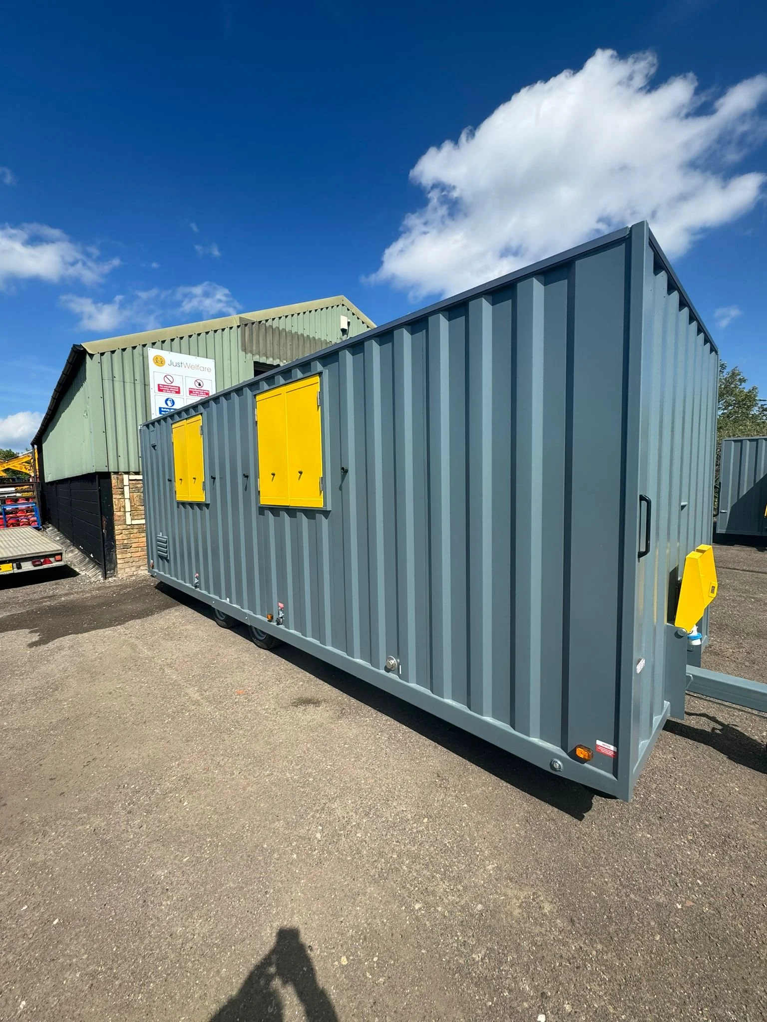 A large gray shipping container with yellow doors and handles, situated outdoors on a gravel surface, with a green building in the background and a blue sky with some clouds.
