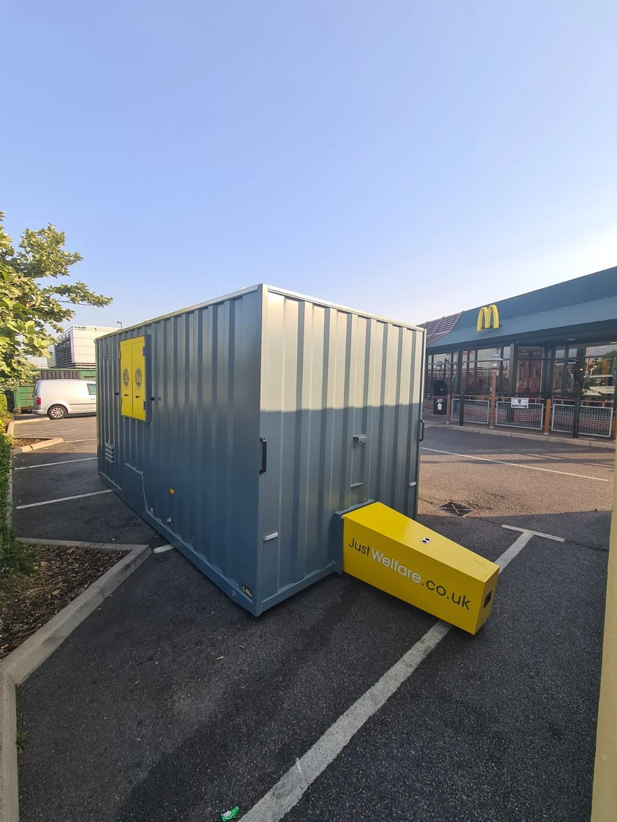 A large metallic container with yellow branding, placed in a parking lot near a McDonald's restaurant. The container is labeled "JustWelfare.co.uk" and has a yellow extension. The sky is clear and the scene appears to be during daylight.