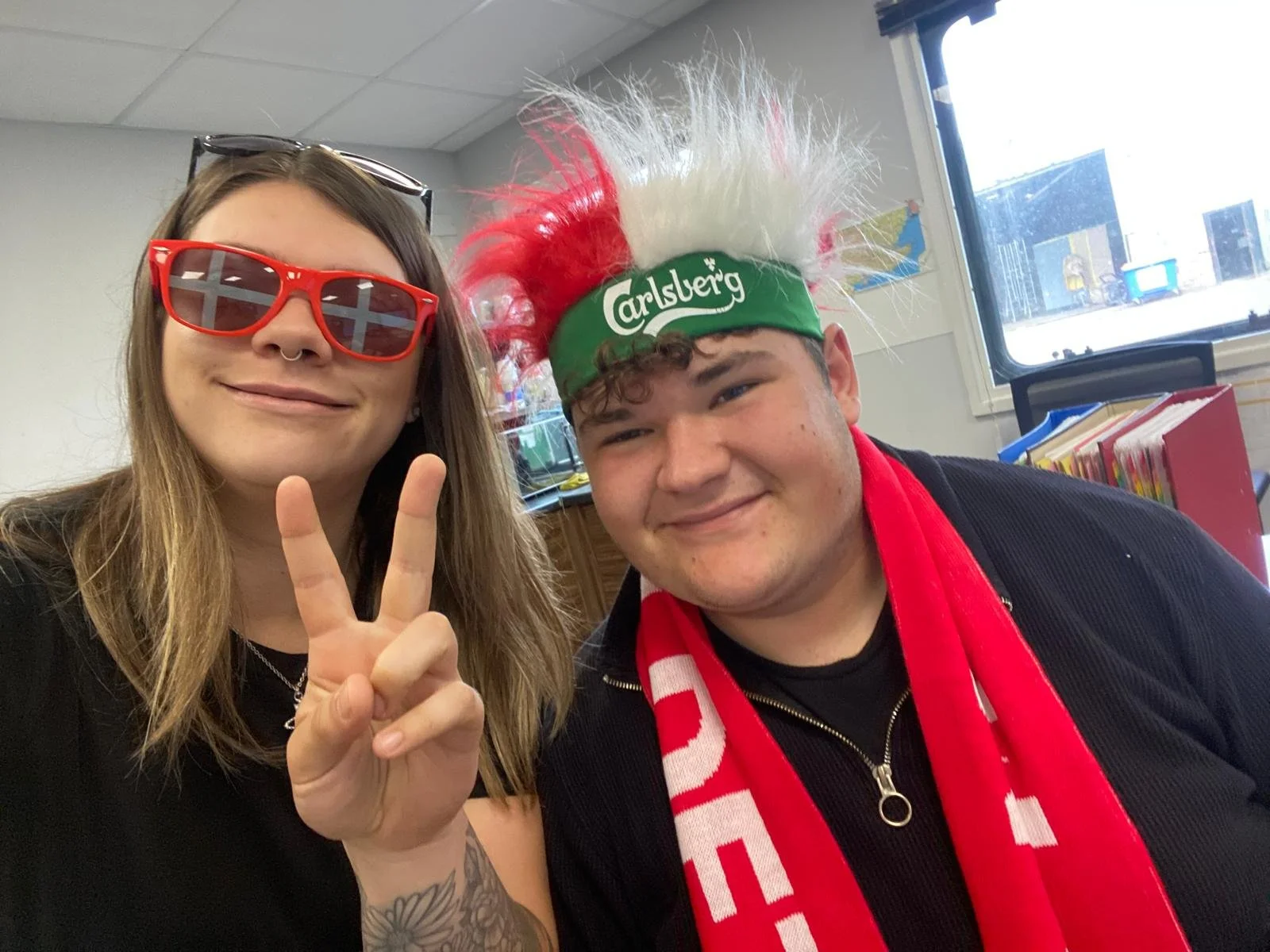 Two people indoors, one making a peace sign, wearing red sunglasses and a septum piercing, the other wearing a green headband with Carlsberg logo and a wig with red and white spiky hair, smiling.