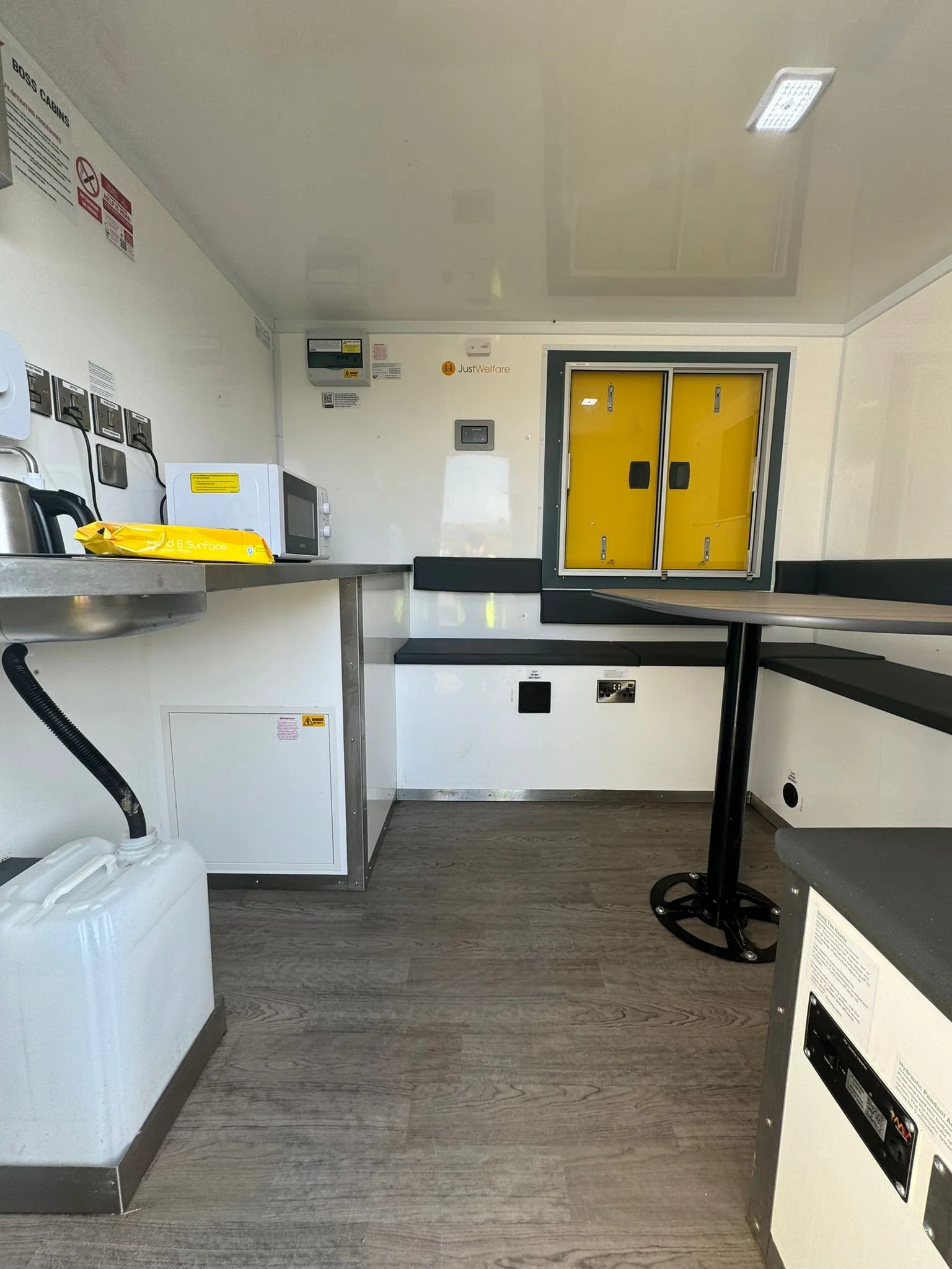 Interior of a clean utility or break room with a gray counter, microwave, electric kettle, yellow emergency box, black bench, and a window with yellow doors. The room has wood-like flooring and white walls with electrical outlets.