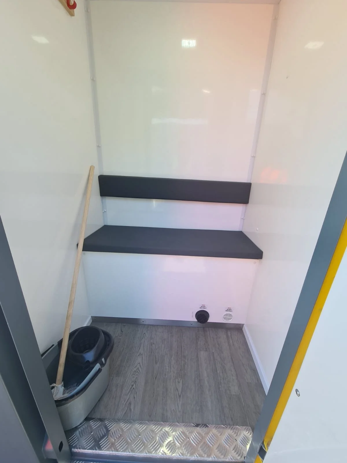 Inside a clean food truck or mobile kitchen space with a small black cushion bench attached to the white wall, a mop and bucket in the corner, and metallic flooring with a diamond plate pattern.