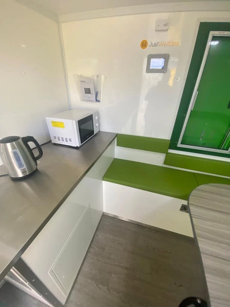 Small kitchenette with a gray countertop, a white microwave, electric kettle, a wall-mounted digital device, green seating area, and a green door or window.