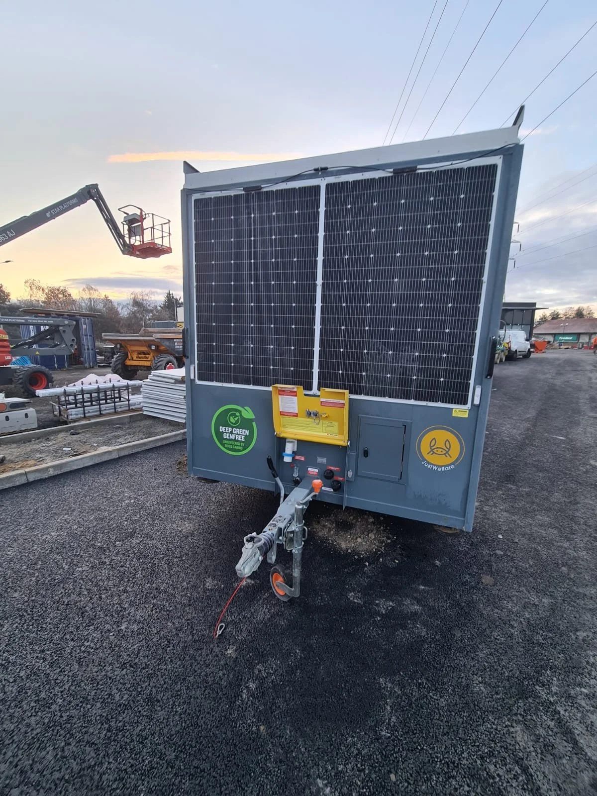 A mobile solar power unit with two large solar panels, set up on a gravel lot, with construction equipment and vehicles in the background at sunset.