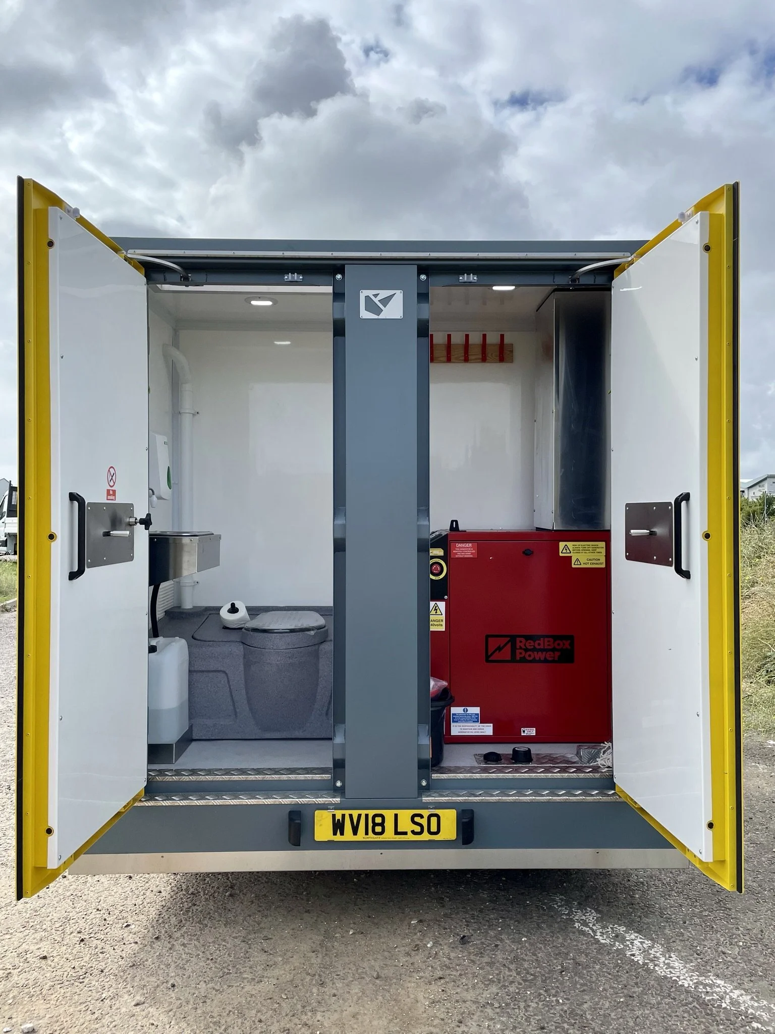 Interior of a mobile restroom unit with a toilet, sink, and a red power generator labeled 'RedBox Power' inside a windowed enclosure with open doors, license plate WV18 LSO, and cloudy sky outside.