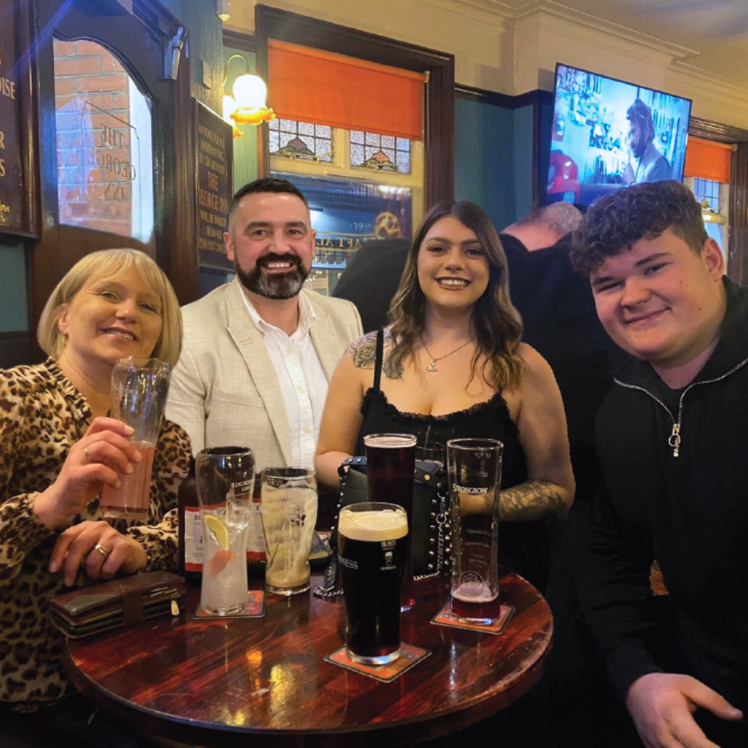 Four people smiling at a pub table with drinks, including beer and cocktails, in a cozy pub with stained glass windows and a TV in the background.