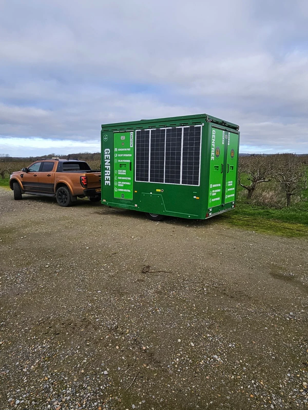 A green portable generator trailer labeled 'Genfire' attached to an orange pickup truck parked on a gravel field with some bare trees and cloudy sky in the background.