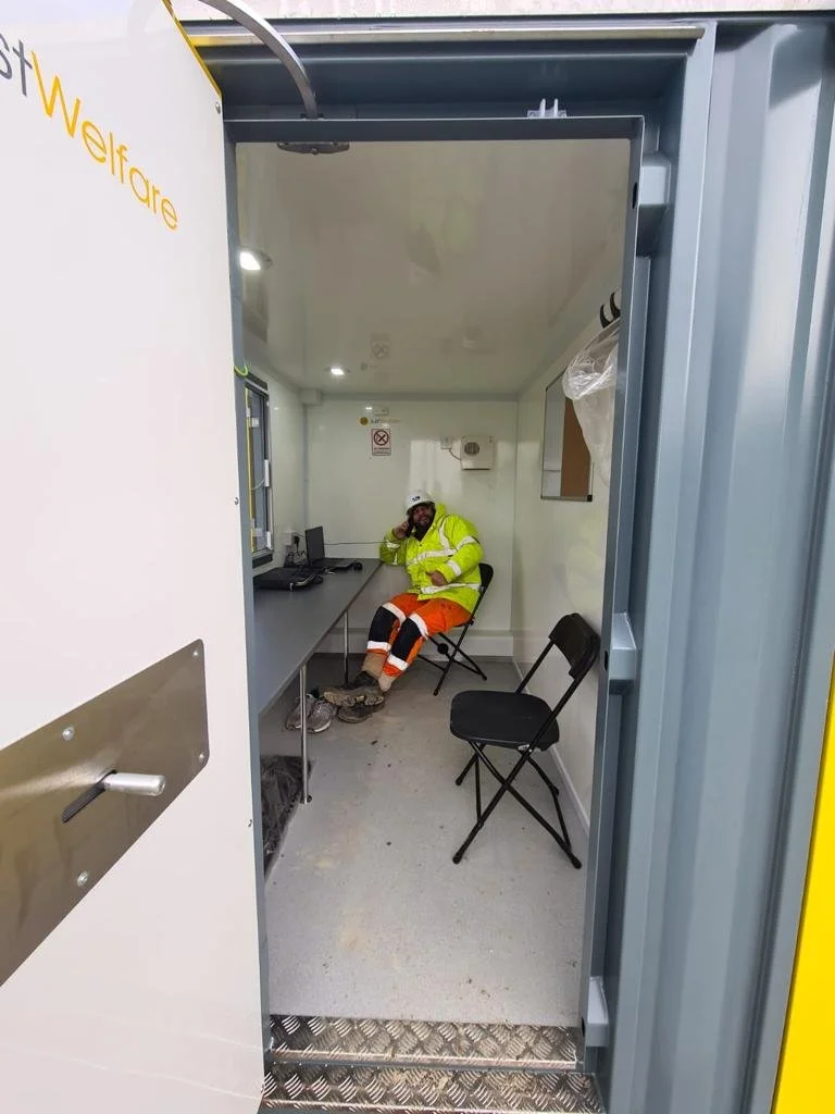 A man in a high-visibility yellow jacket and orange pants sits inside a small, enclosed booth, talking on a cellphone. There are two black chairs, a desk with a computer, and a window or opening on the wall.