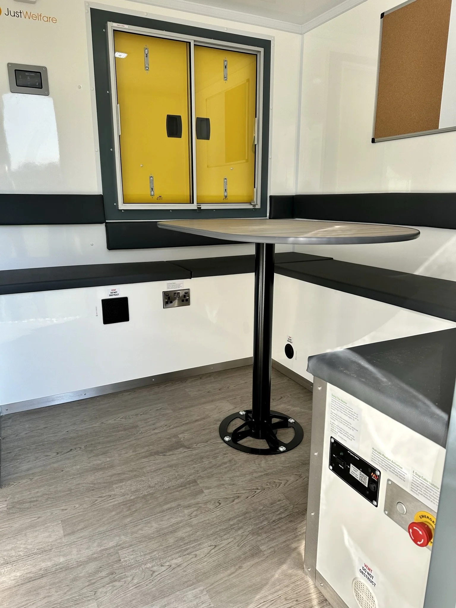 Interior of a room with a yellow lift up window, black cushioned bench seat, a small round table, and electrical outlets.