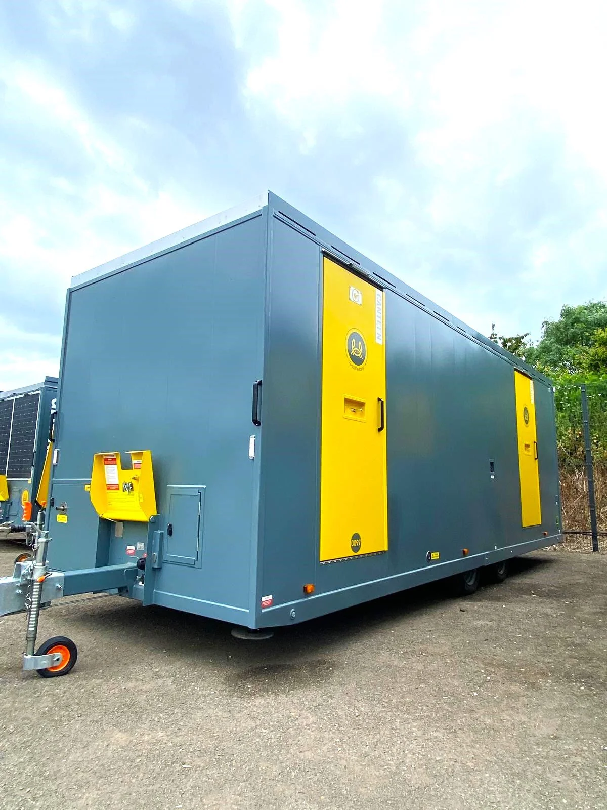 Large blue and yellow mobile generator or electrical equipment container outdoors on dirt ground with trees and sky in the background.