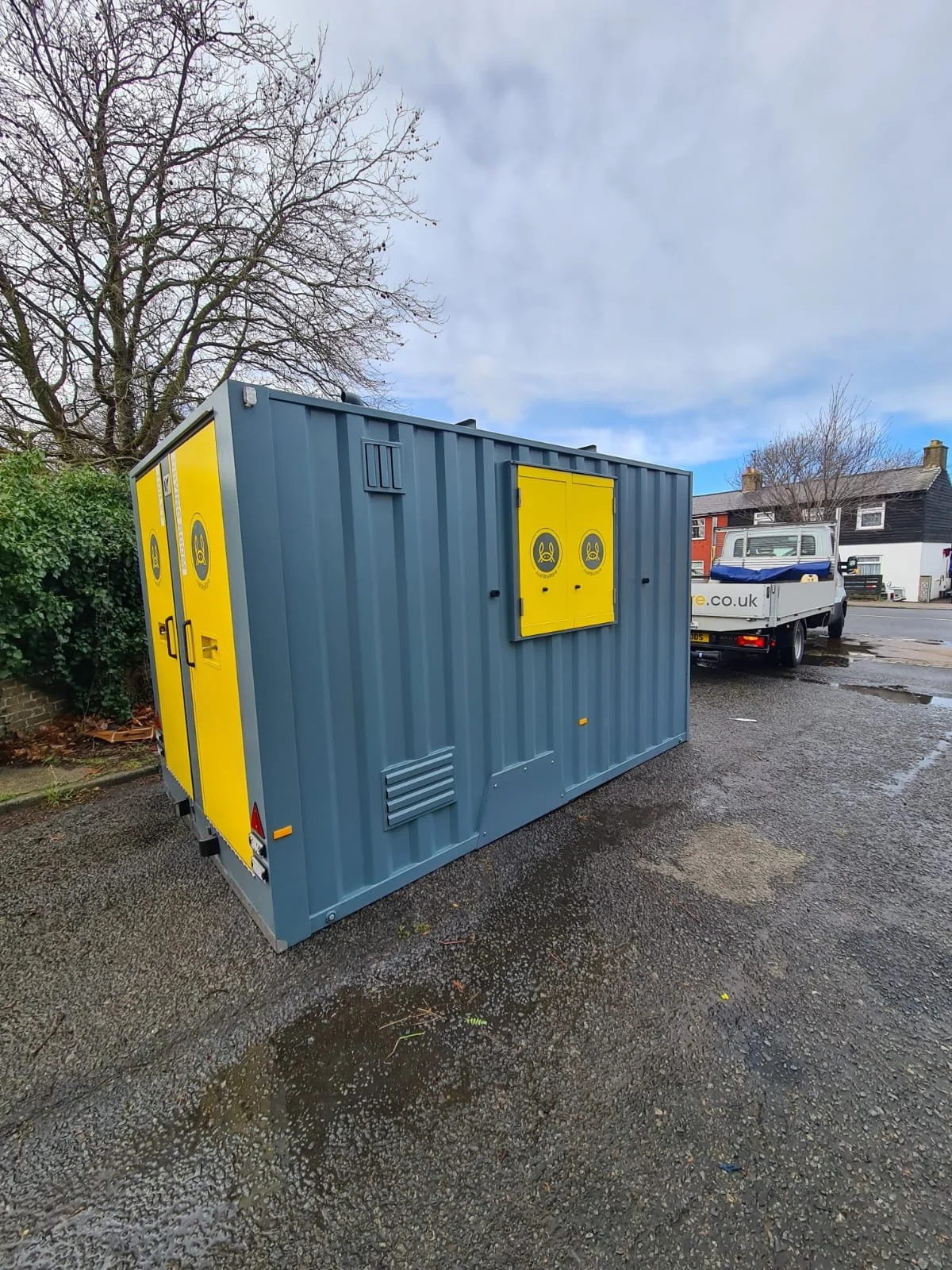 A blue and yellow mobile electrical utility box on a gravel surface, with a tree and residential buildings in the background.