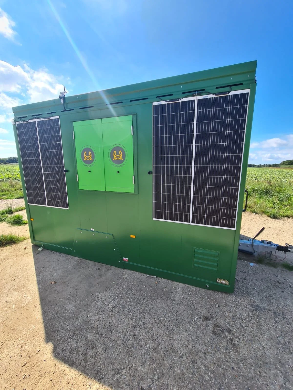 A green portable energy or electrical unit with solar panels on the side, located outdoors in a field under a blue sky.