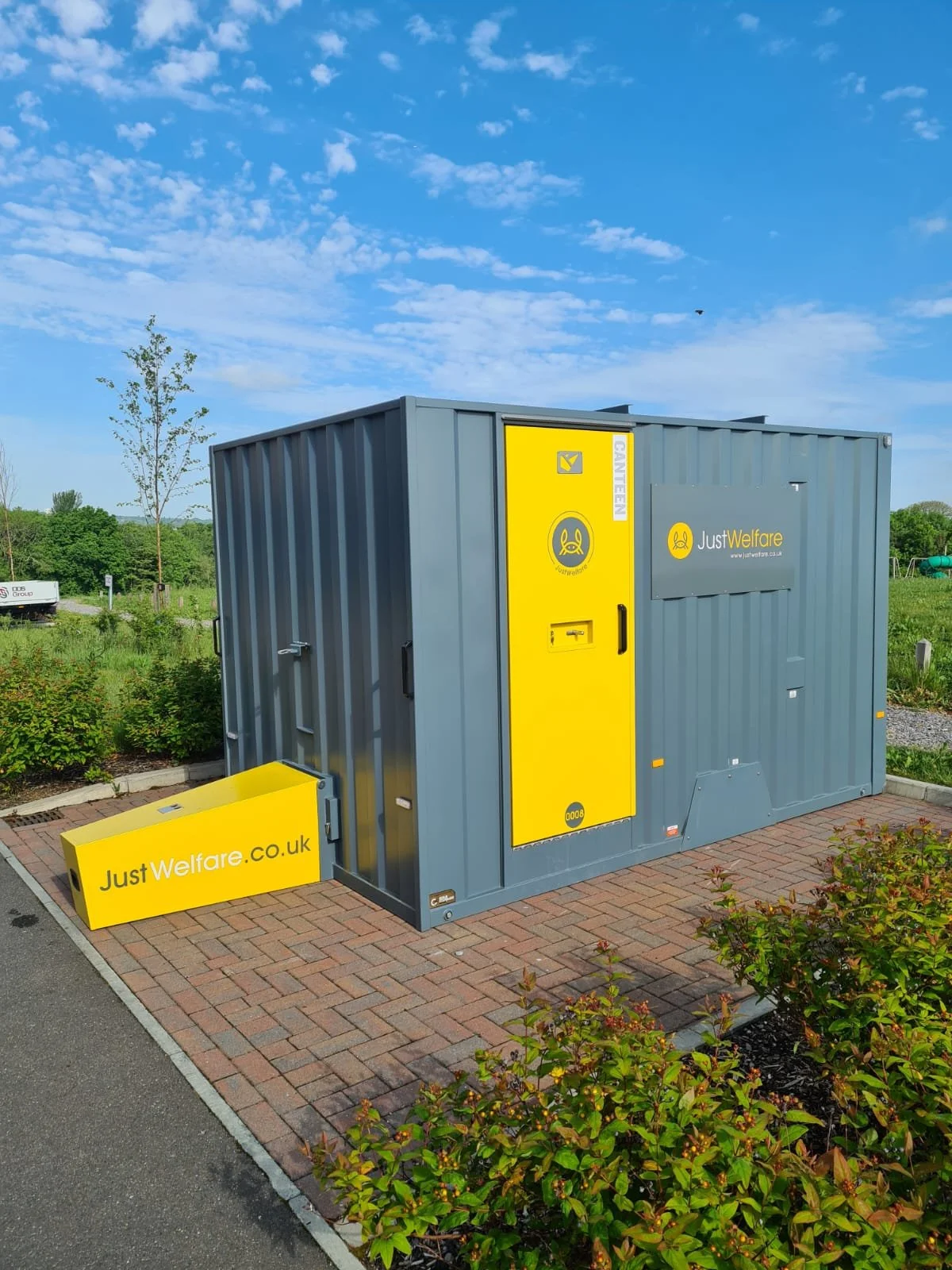 A portable welfare and food donation container with yellow and gray coloring, situated outdoors on a brick-paved area with greenery and trees in the background under a blue sky with scattered clouds.