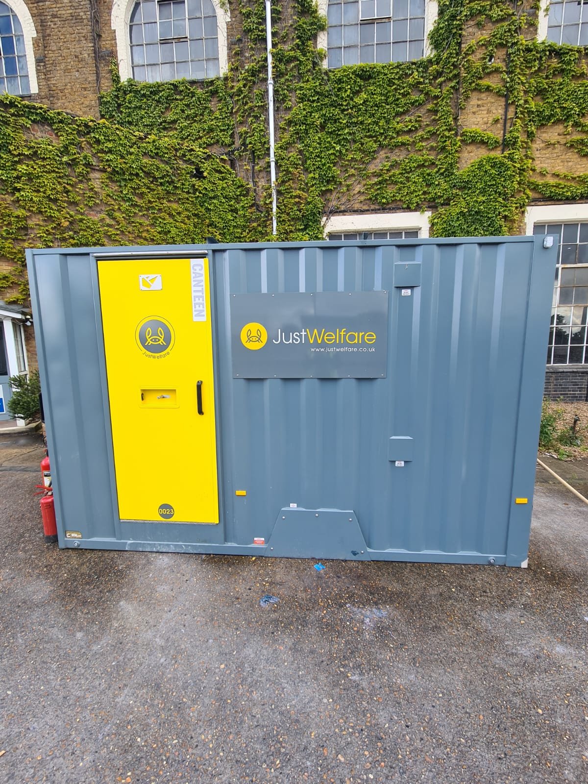 A metal container with 'JustWelfare' branding, featuring a yellow door labeled 'CANTEN' and a logo, situated outdoors against a background of a brick building covered with green ivy.