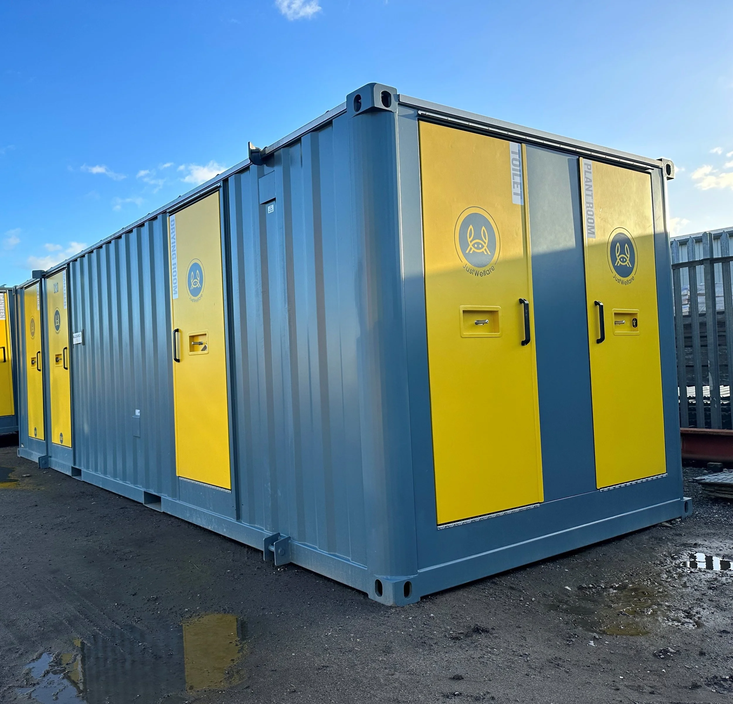 A large gray and yellow portable restroom trailer with multiple doors, set outdoors on a dirt surface with a blue sky background.