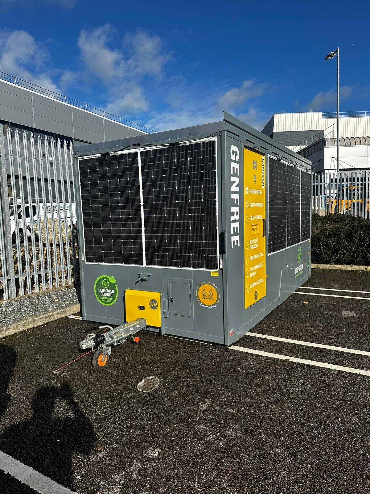 A portable generator with solar panels installed on top, parked in a designated parking space outdoors near a fence and a large building, under a partly cloudy sky.