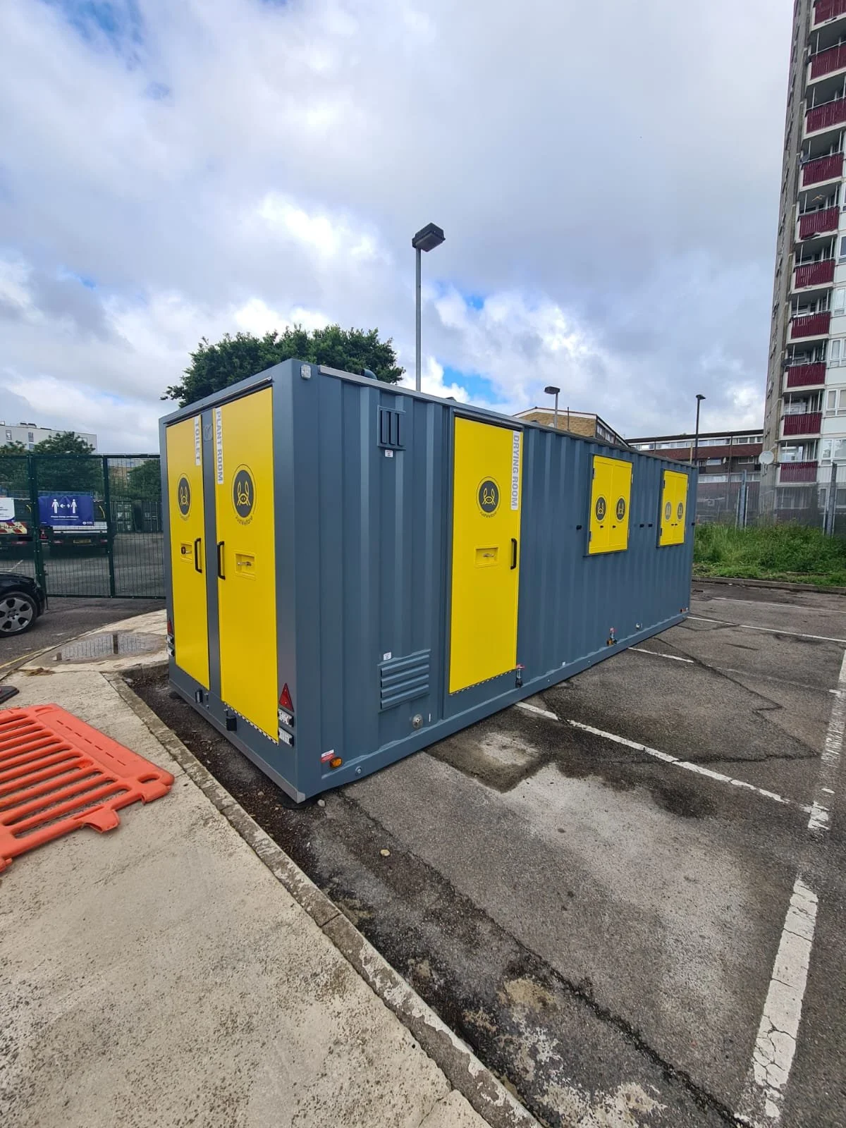 A large blue container with yellow warnings and doors outdoor in a parking lot, with a tall building and cloudy sky in the background.