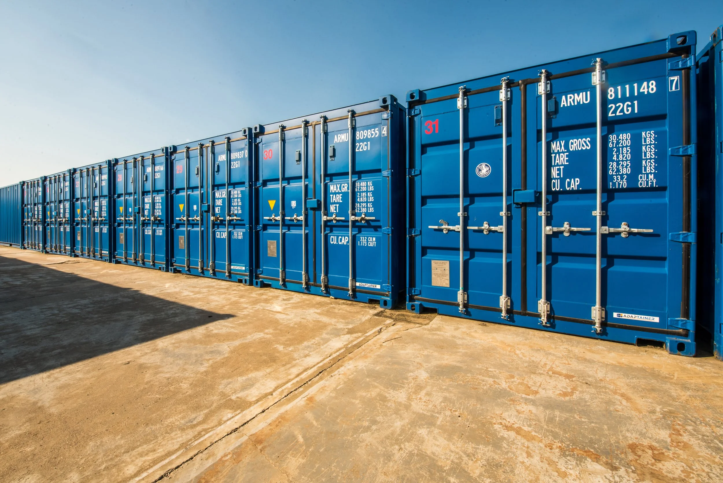 A row of blue shipping containers lined up on a concrete lot under a clear blue sky.
