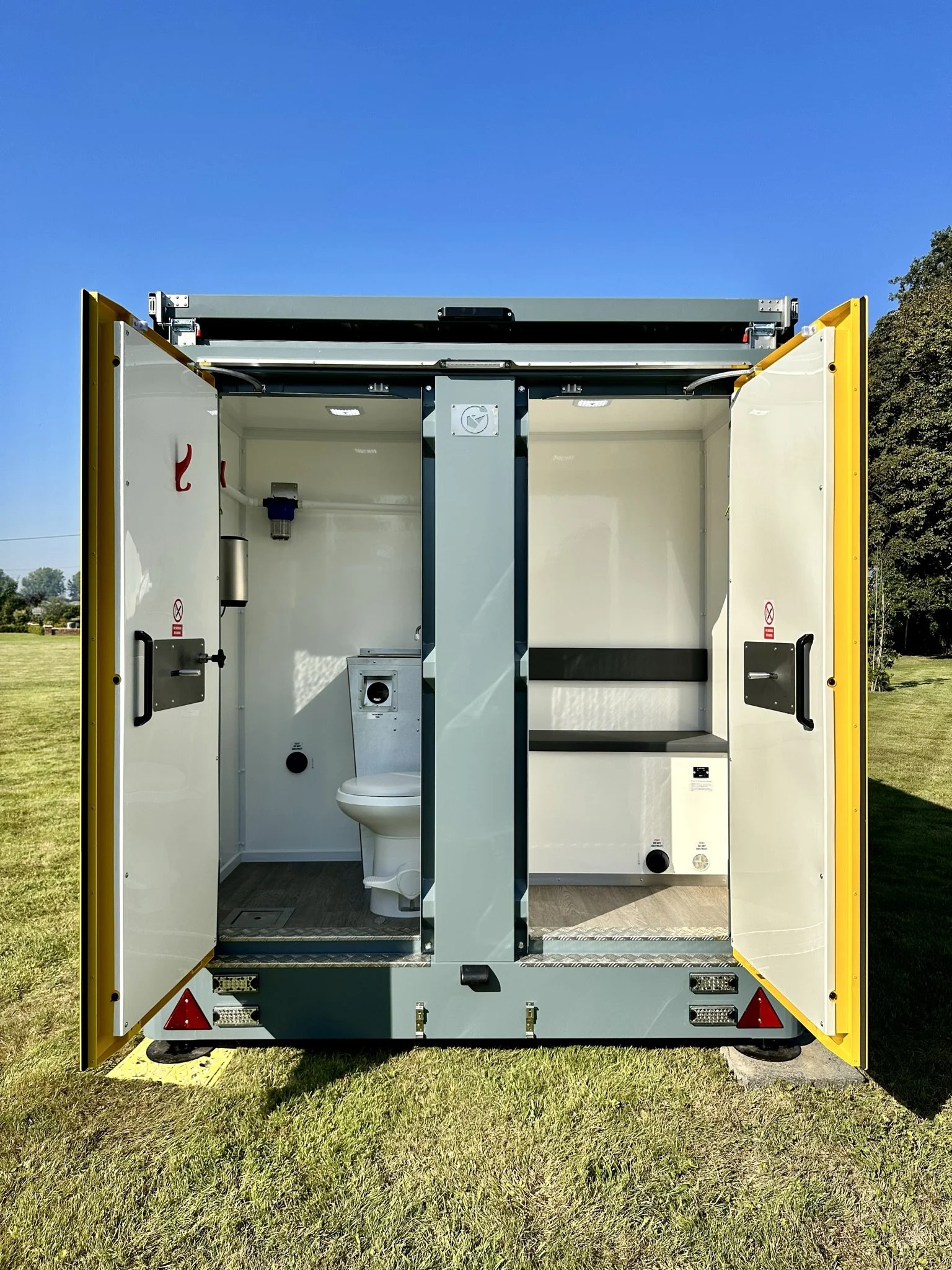 A portable mobile bathroom unit with two separate compartments, one featuring a toilet and the other a bench and storage space. The doors are open, and the unit is situated outdoors on a grassy field with a clear blue sky.