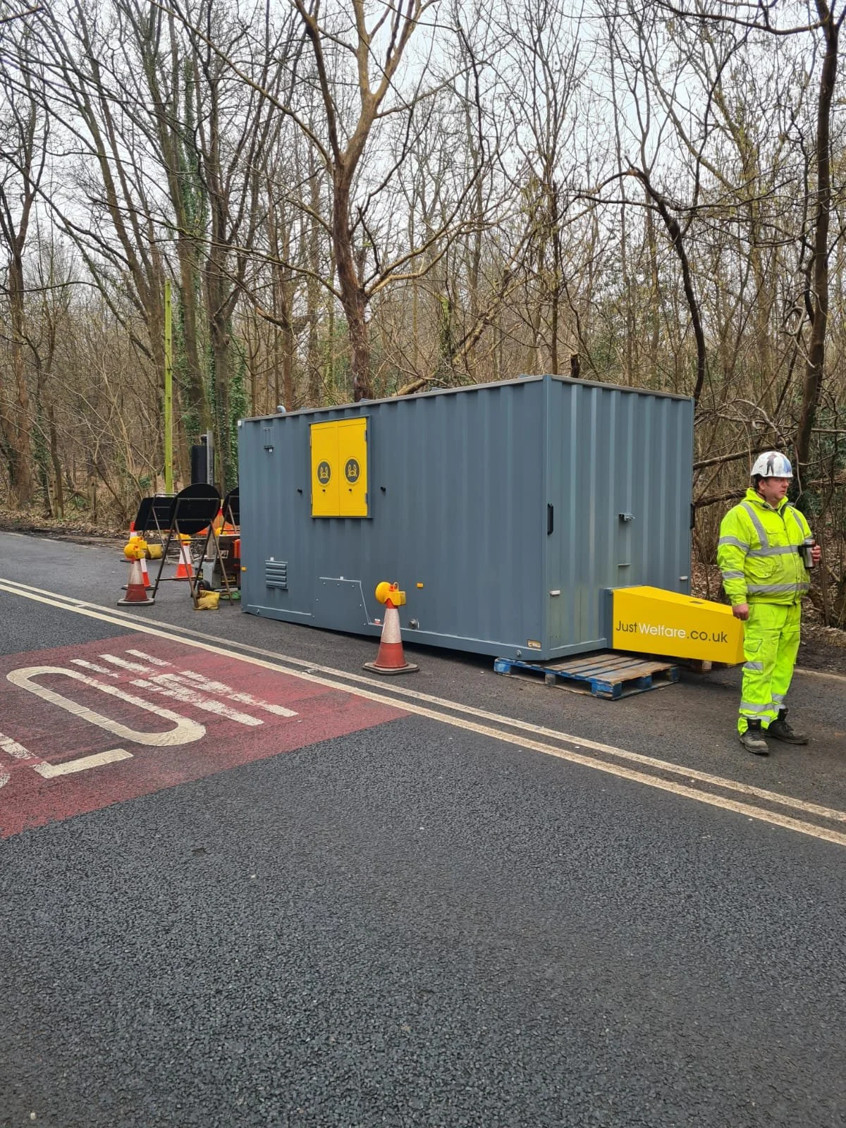 A construction worker in a yellow reflective suit and helmet standing next to a portable unit with a sign that reads 'JustWelfare.co.uk' on a roadside with traffic cones, a stop sign painted on the road, and leafless trees in the background.