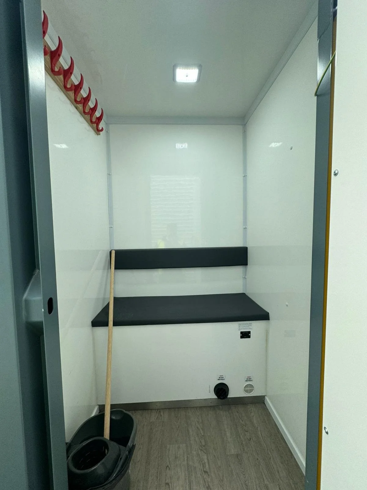 Empty medical or public utility booth with a black padded bench, cleaning supplies, and hooks on the wall.