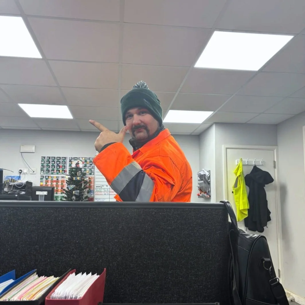 Man in construction attire and a knit hat giving a finger gun pose while standing in an office or break room.