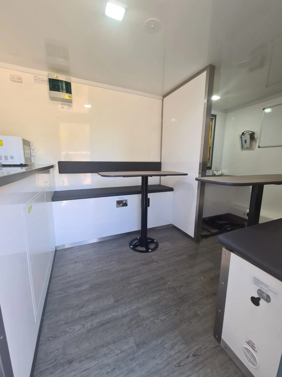 Interior of a small room with a white wall, a small black table, and a black bench. There are appliances, an electrical outlet, and a whiteboard on the walls.