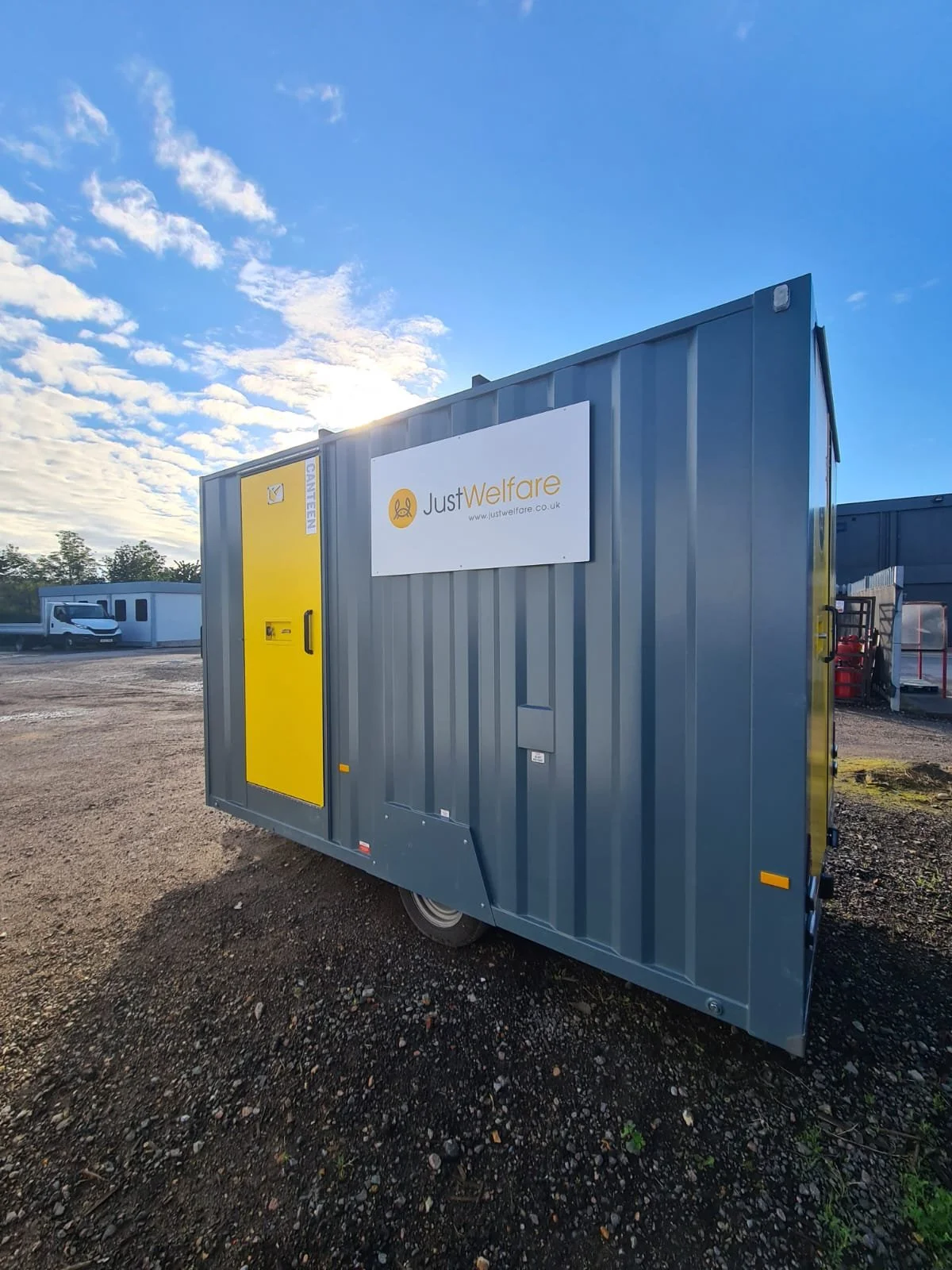A portable welfare unit with a yellow door, parked outdoors on a gravel lot, with a sign reading "Just Welfare" above it, under a partially cloudy sky.