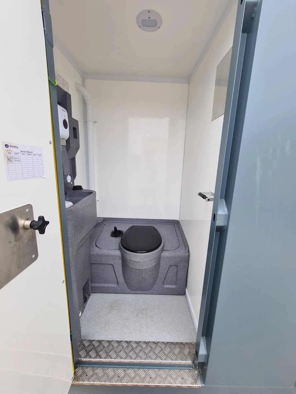 Small enclosed portable bathroom with gray raised toilet seat, sink, hand sanitizer dispenser, and a small mirror. The interior is minimal and functional, with white walls and a metallic step at the entrance.