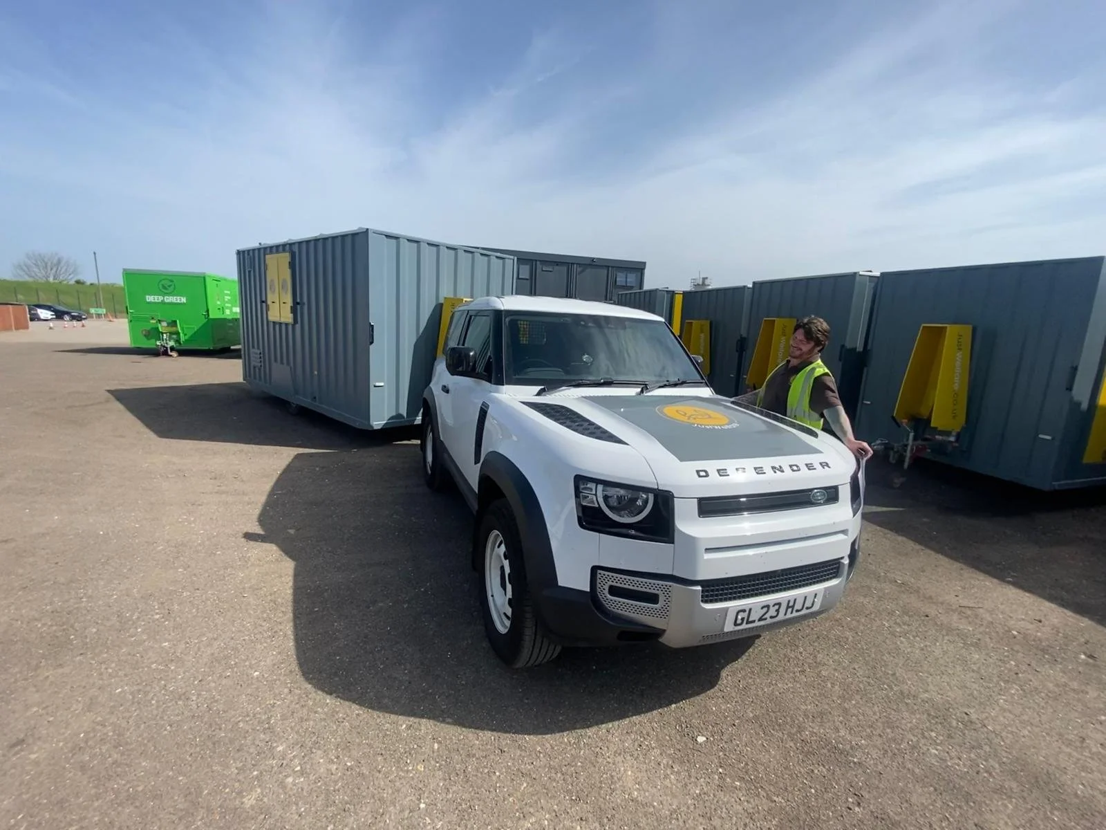White autonomous service robot vehicle with the word 'DEFENDER' on the front, parked in a lot next to delivery containers. A person with a safety vest is standing nearby, smiling.