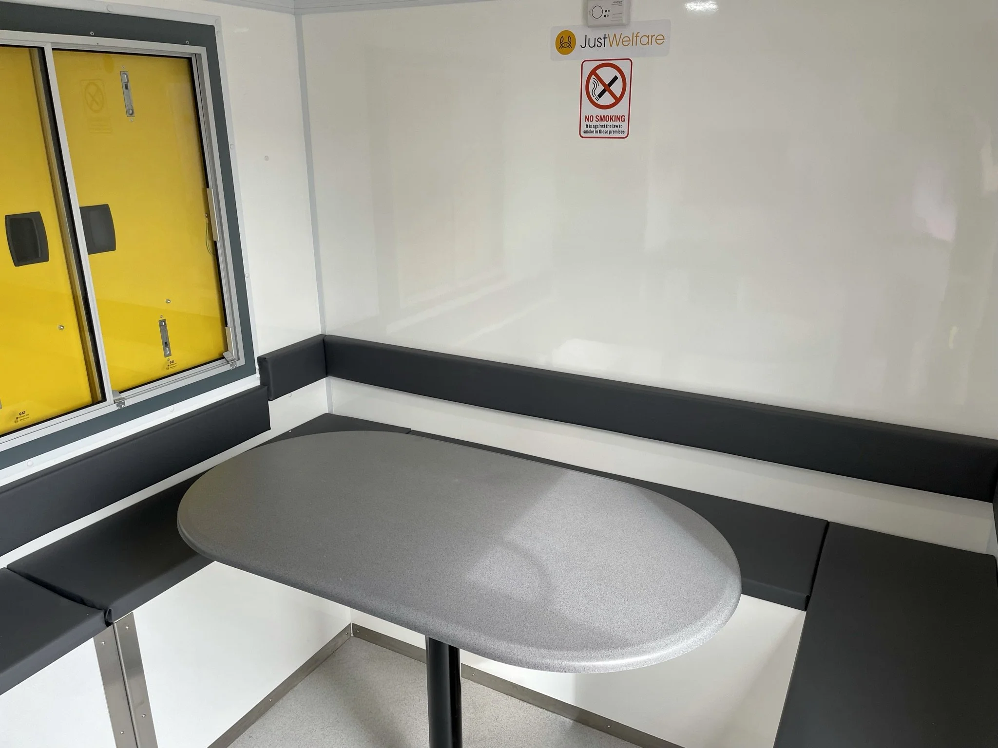 Empty corner inside a food pickup area with yellow lockers, a round gray table, and a white wall with a 'JustWelfare' sign and a no smoking sign.