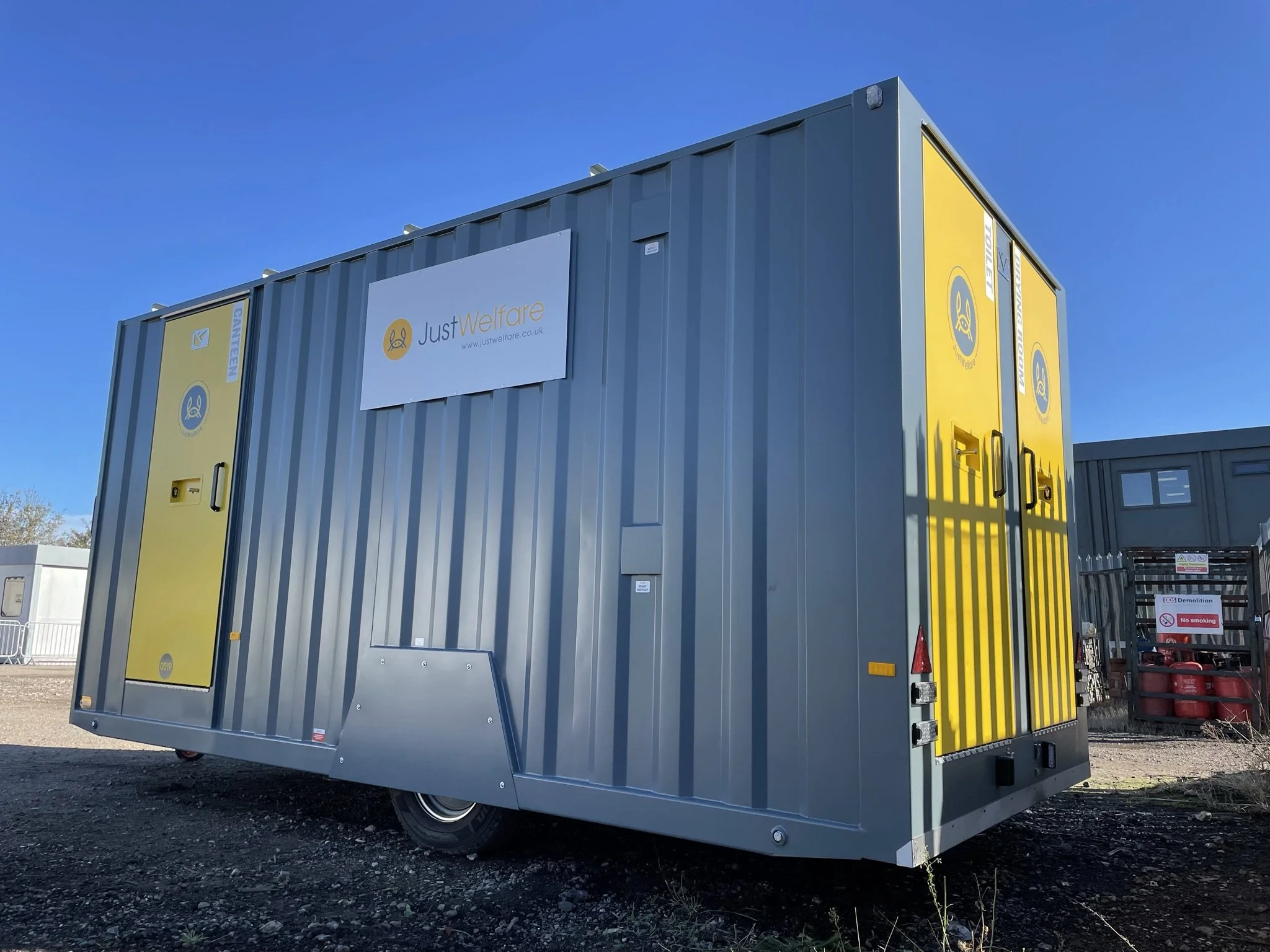 A large gray container with yellow doors, labeled 'Just Welfare' and decorated with a blue and yellow logo, sitting outdoors on a gravel surface with a clear blue sky overhead.