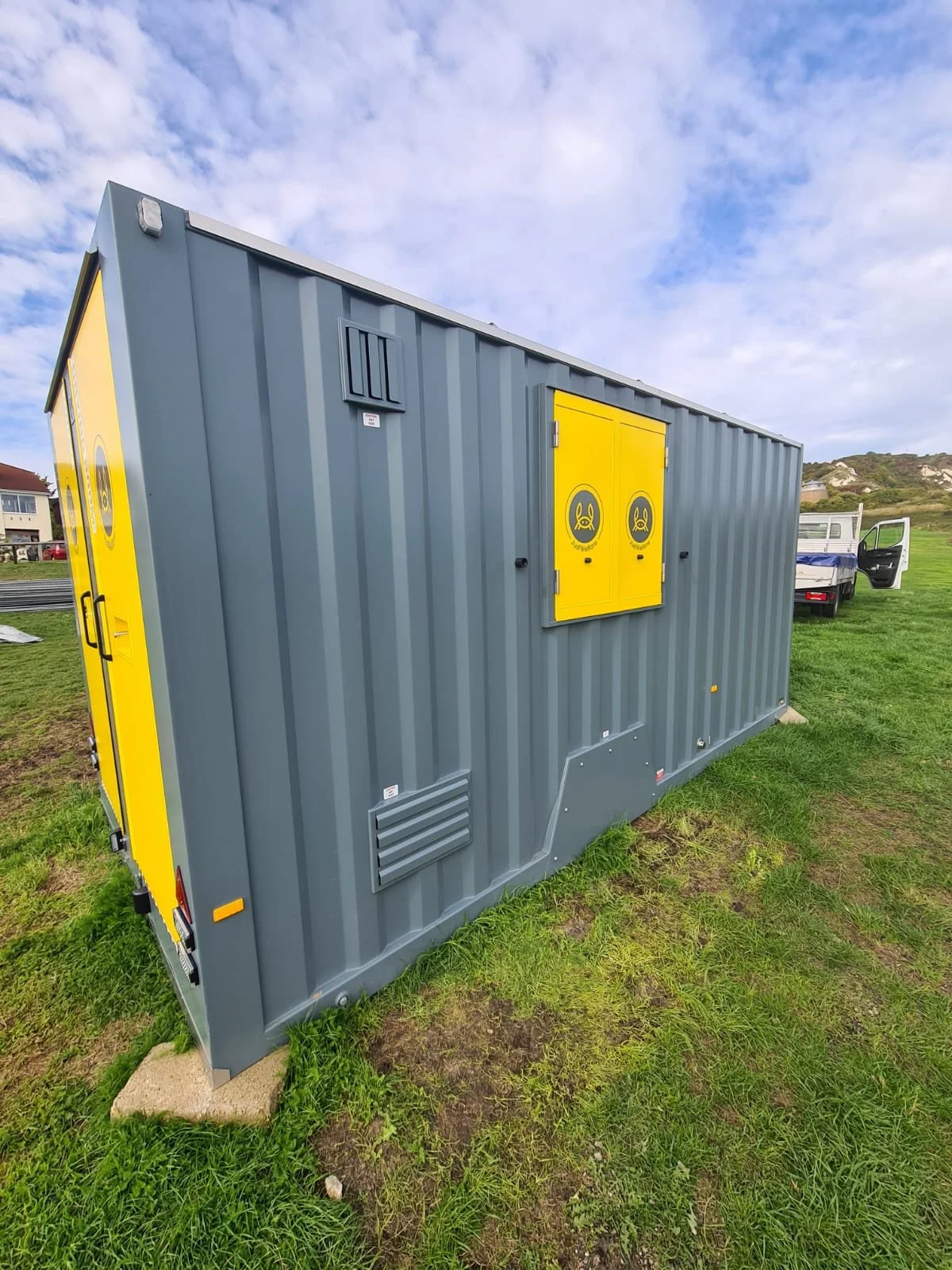 A large gray utility or storage container with yellow doors and logos, situated outdoors on a grassy area under a cloudy sky, with a truck and some residential buildings in the background.