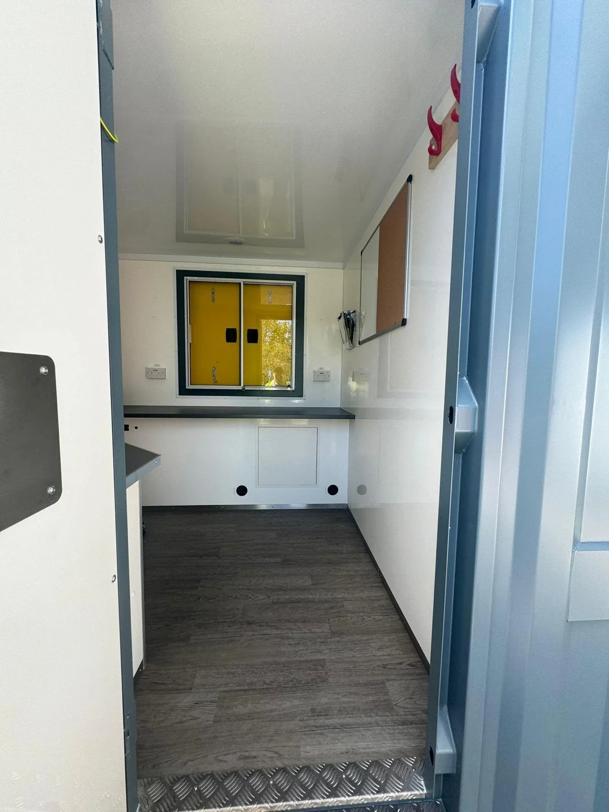 Empty, clean mobile office or trailer interior with small window, white walls, gray countertop, cork board, and wood flooring.