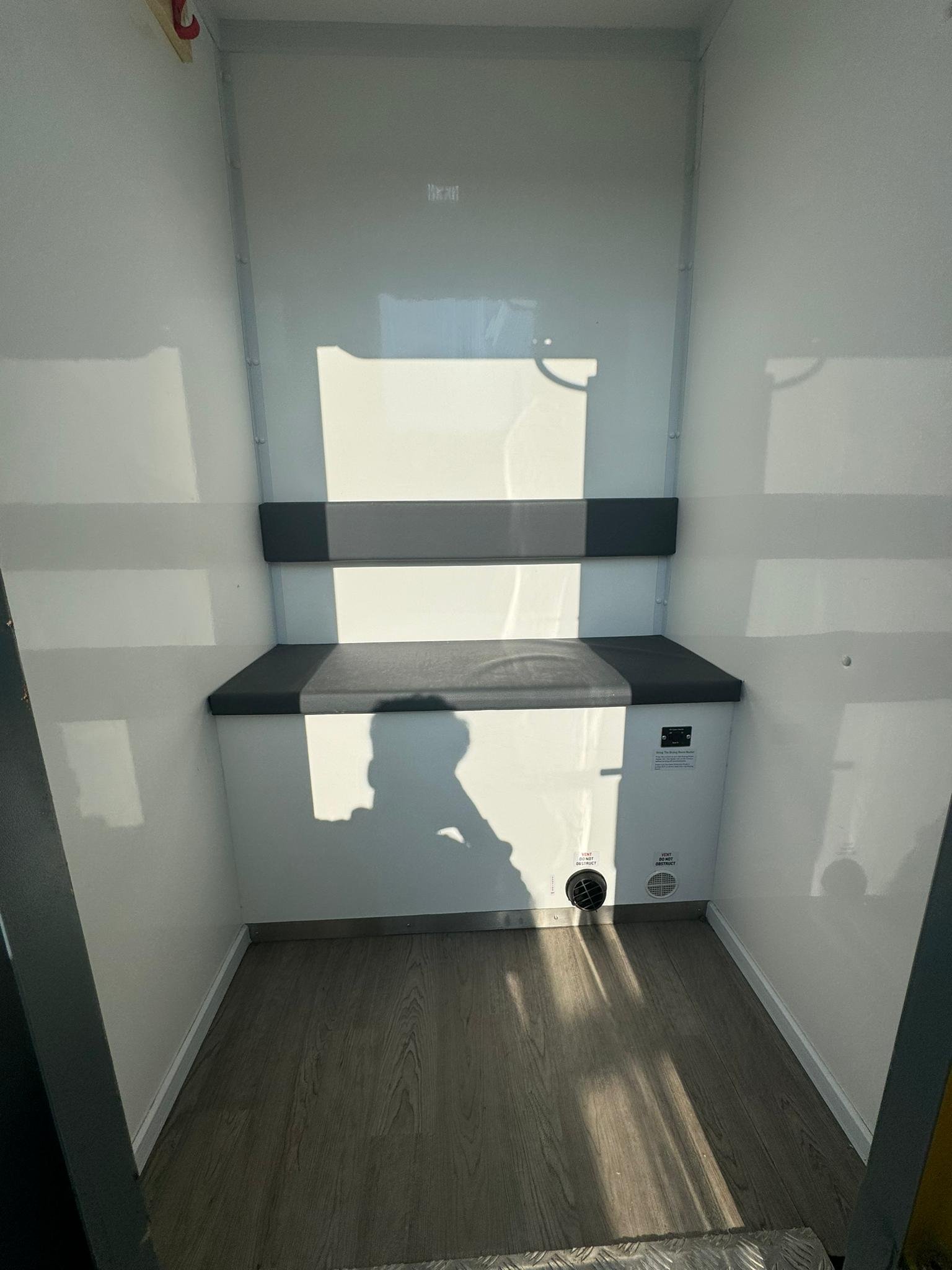 Empty enclosed space with white walls, a black cushioned bench on the back wall, and a vent on the lower right side.