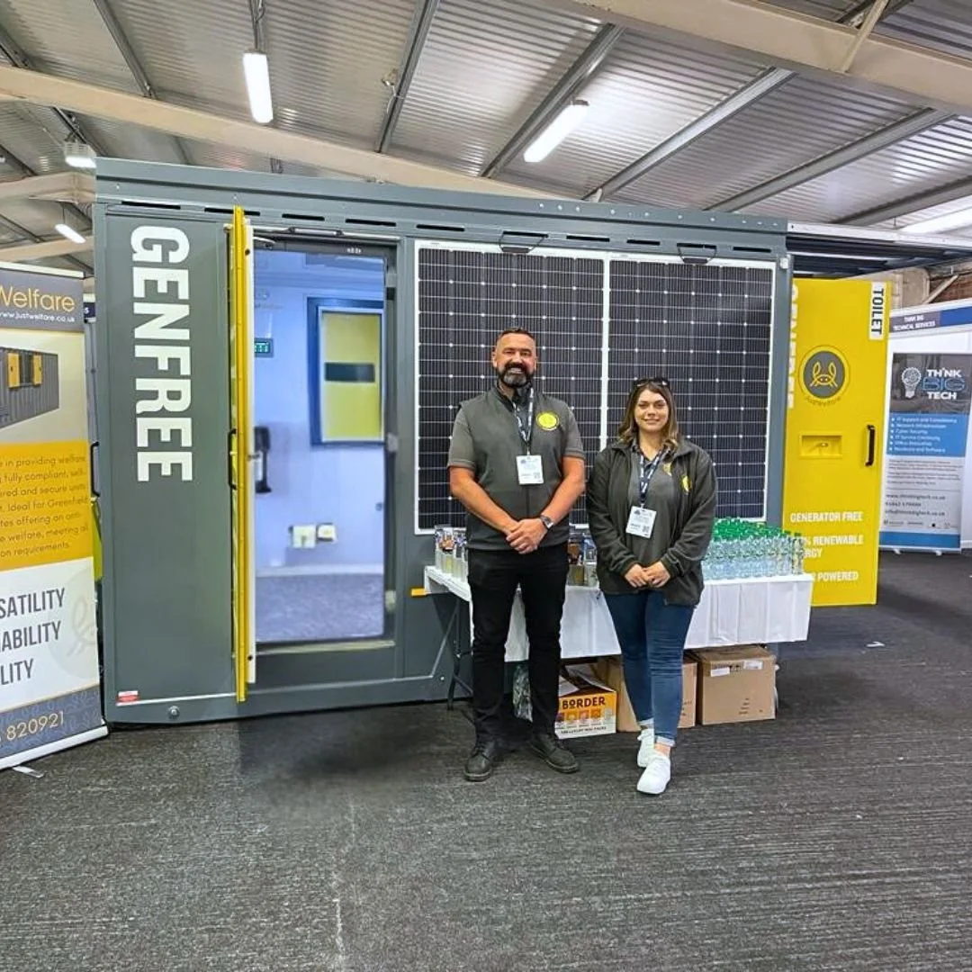 Two people standing in front of a large mobile solar power generator with solar panels, inside a convention hall.